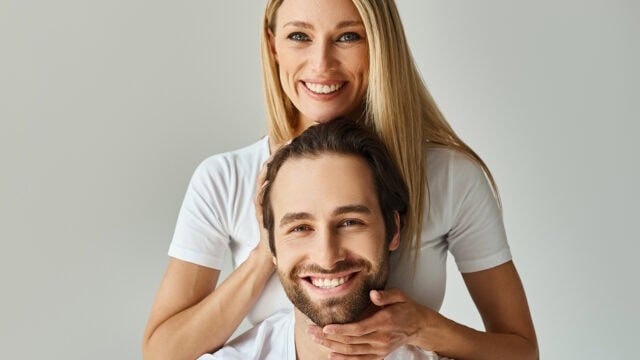 A smiling woman with blonde hair standing behind a smiling man, resting her hands on his head and chin.