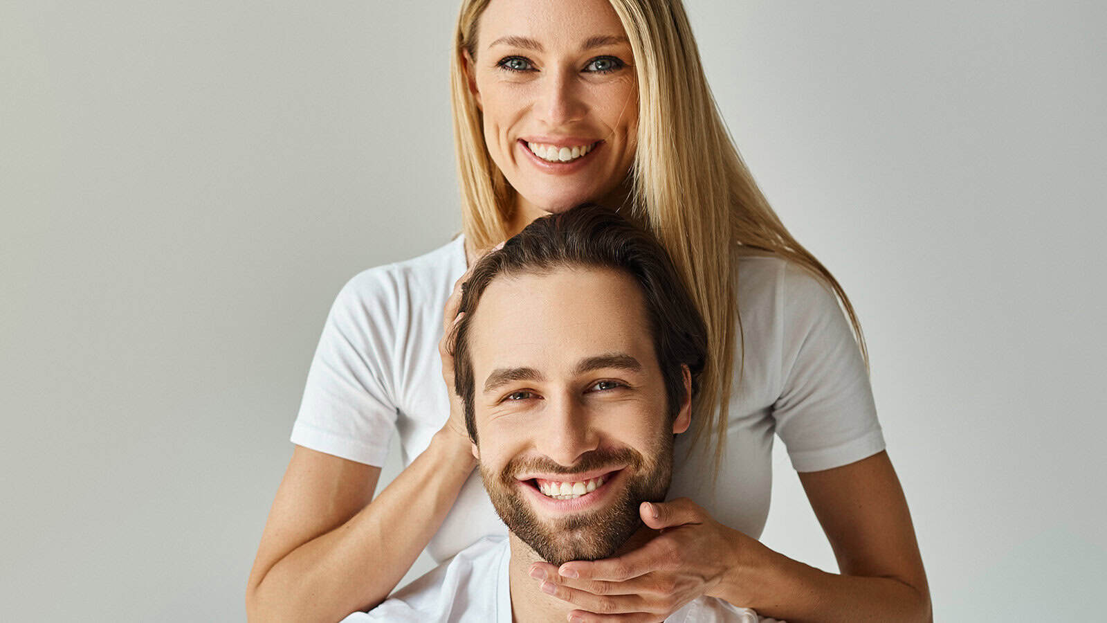 A smiling woman with blonde hair standing behind a smiling man, resting her hands on his head and chin.