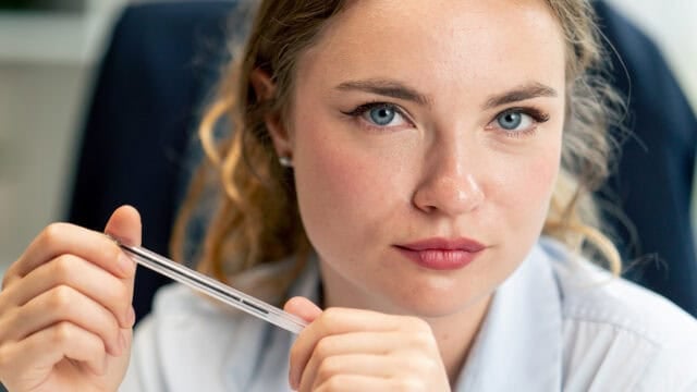 A woman with blue eyes and curly blonde hair looks directly at the camera, holding a pen in both hands. She wears a light-colored shirt and sits in a professional setting.