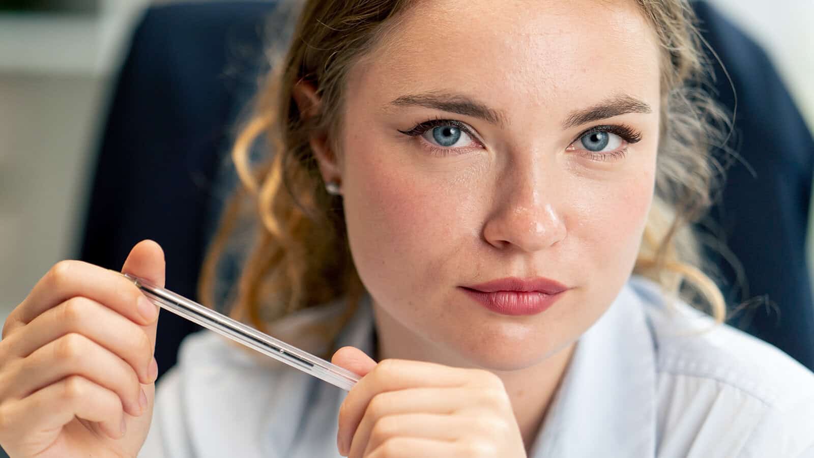 A woman with blue eyes and curly blonde hair looks directly at the camera, holding a pen in both hands. She wears a light-colored shirt and sits in a professional setting.