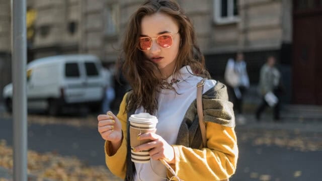 A young woman in a yellow coat and pink-tinted sunglasses holds a paper coffee cup while standing on a city street.