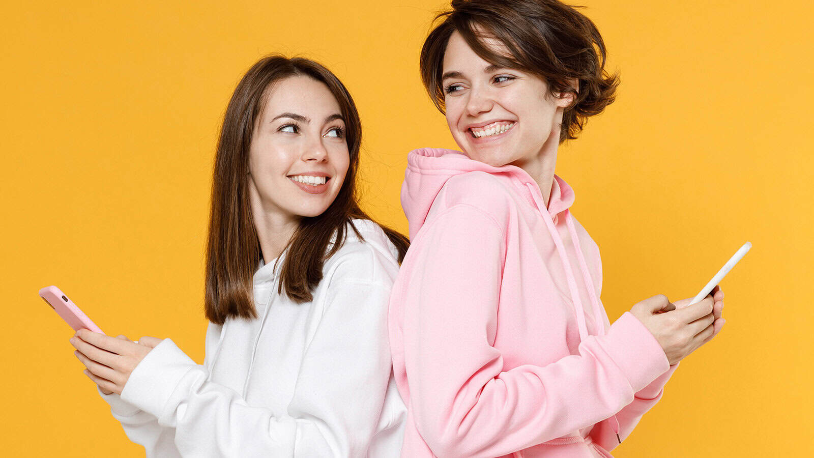 Two smiling women in hoodies stand back-to-back, looking at each other while holding smartphones against a yellow background.