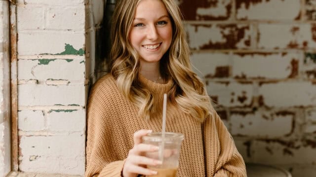 A smiling young woman in a tan sweater holding a plastic cup with a straw.