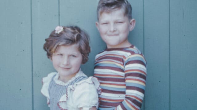 A young girl and boy standing side-by-side against a green paneled wall.