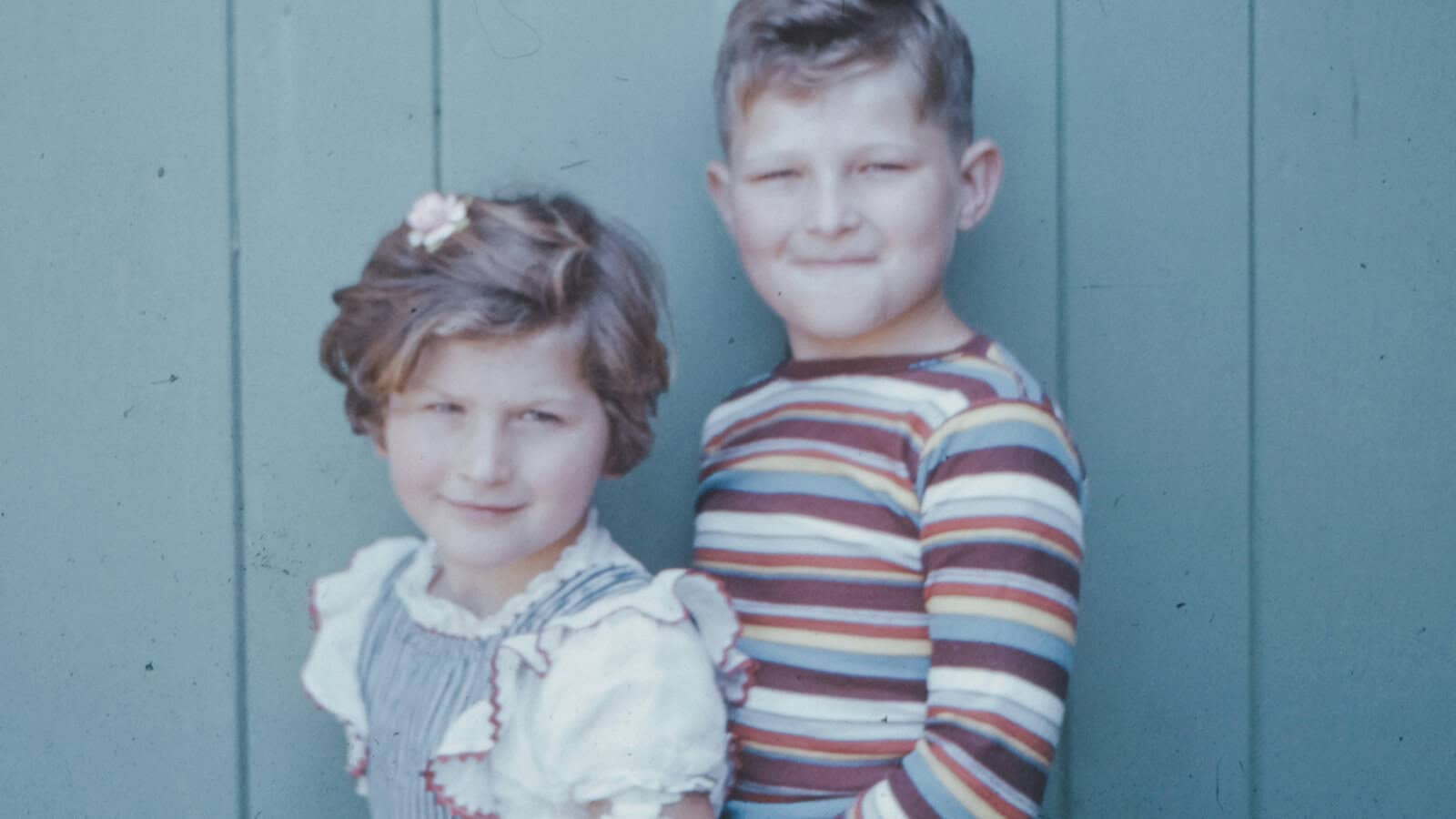 A young girl and boy standing side-by-side against a green paneled wall.