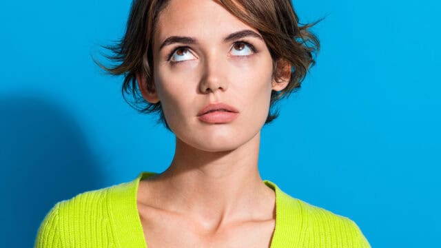 Woman with short brown hair wearing a bright yellow-green sweater stands against a blue background, looking upward with an expression that appears thoughtful or slightly annoyed.
