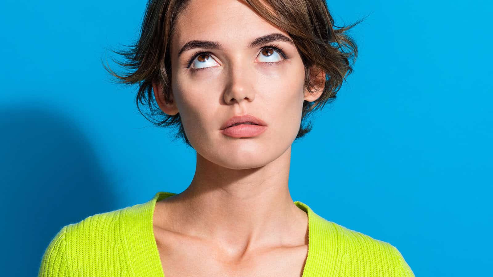 Woman with short brown hair wearing a bright yellow-green sweater stands against a blue background, looking upward with an expression that appears thoughtful or slightly annoyed.