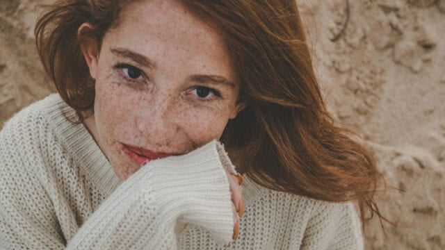 A woman with freckles wearing a cream-colored sweater rests her chin on her hand.