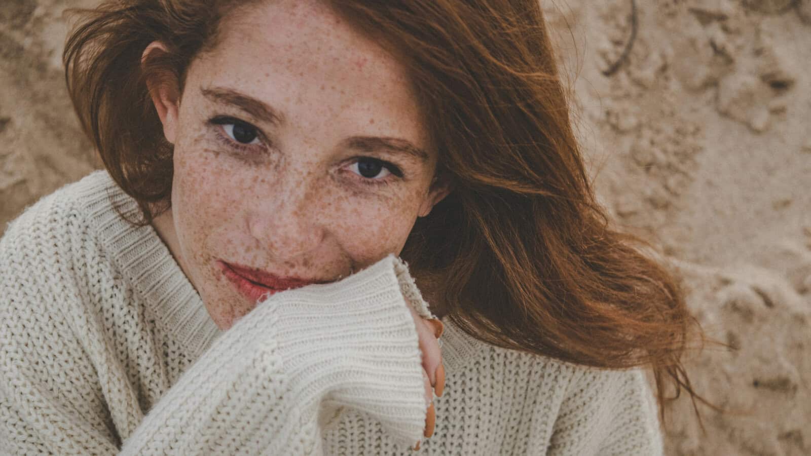 A woman with freckles wearing a cream-colored sweater rests her chin on her hand.