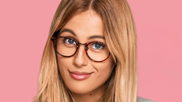 A woman with blonde hair wearing round tortoise-shell glasses against a pink background.