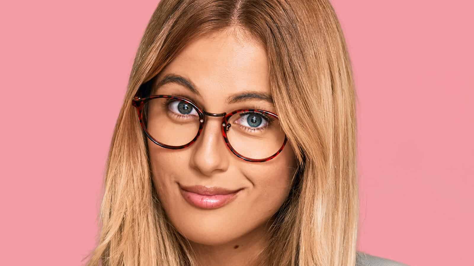 A woman with blonde hair wearing round tortoise-shell glasses against a pink background.