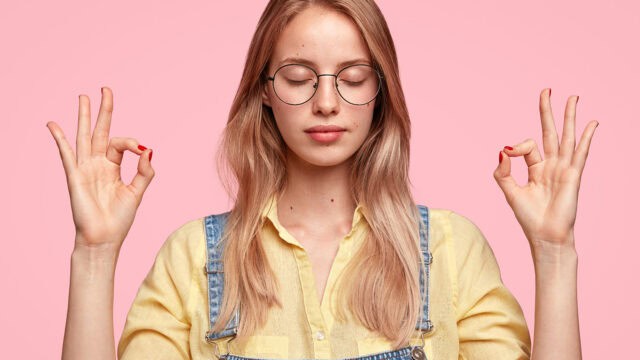 Young woman with long blonde hair, wearing glasses and denim overalls over a yellow shirt, stands against a pink background, eyes closed, holding both hands in a meditation gesture.