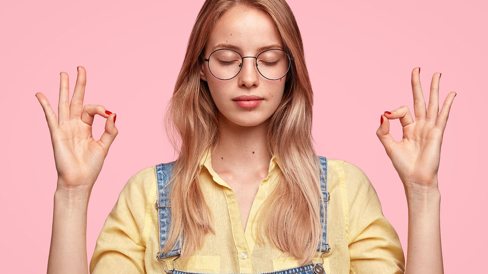 Young woman with long blonde hair, wearing glasses and denim overalls over a yellow shirt, stands against a pink background, eyes closed, holding both hands in a meditation gesture.