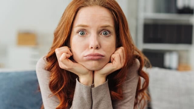 A woman with red hair rests her chin on her hands, cheeks puffed out, and eyes wide open, looking directly at the camera with an expression of boredom or frustration.