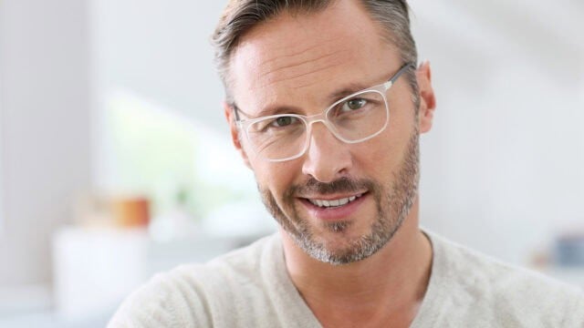 A man with short brown hair, light beard, and clear-framed glasses smiles at the camera. He is wearing a light beige sweater and is indoors with a bright, blurred background.
