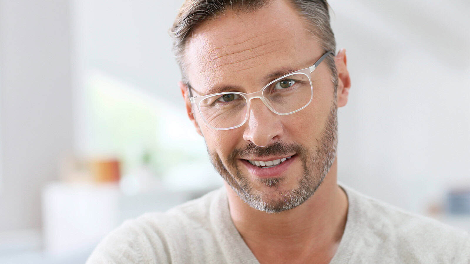A man with short brown hair, light beard, and clear-framed glasses smiles at the camera. He is wearing a light beige sweater and is indoors with a bright, blurred background.