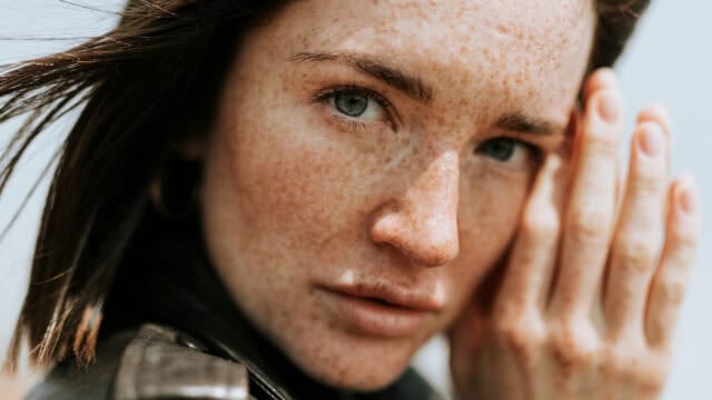 Close-up portrait of a woman with freckles, looking directly at the camera with her hand raised toward her face.