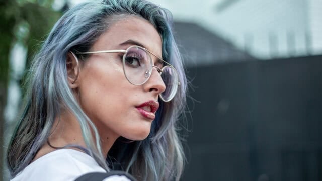 A woman with blue-tinted hair wearing gold-rimmed glasses and glitter on her eyelids and lips.