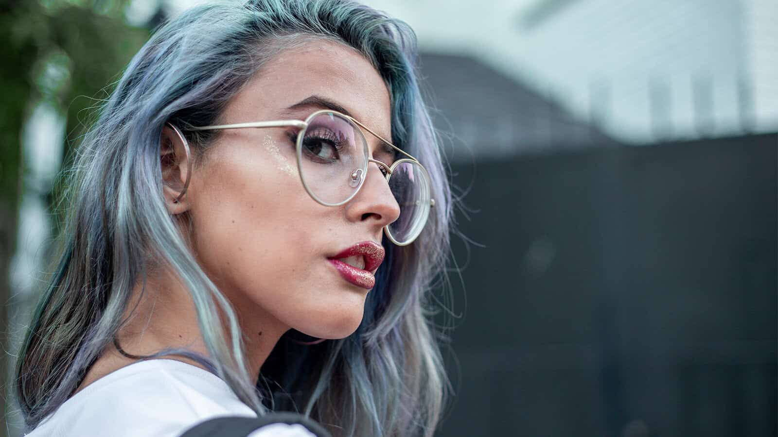 A woman with blue-tinted hair wearing gold-rimmed glasses and glitter on her eyelids and lips.