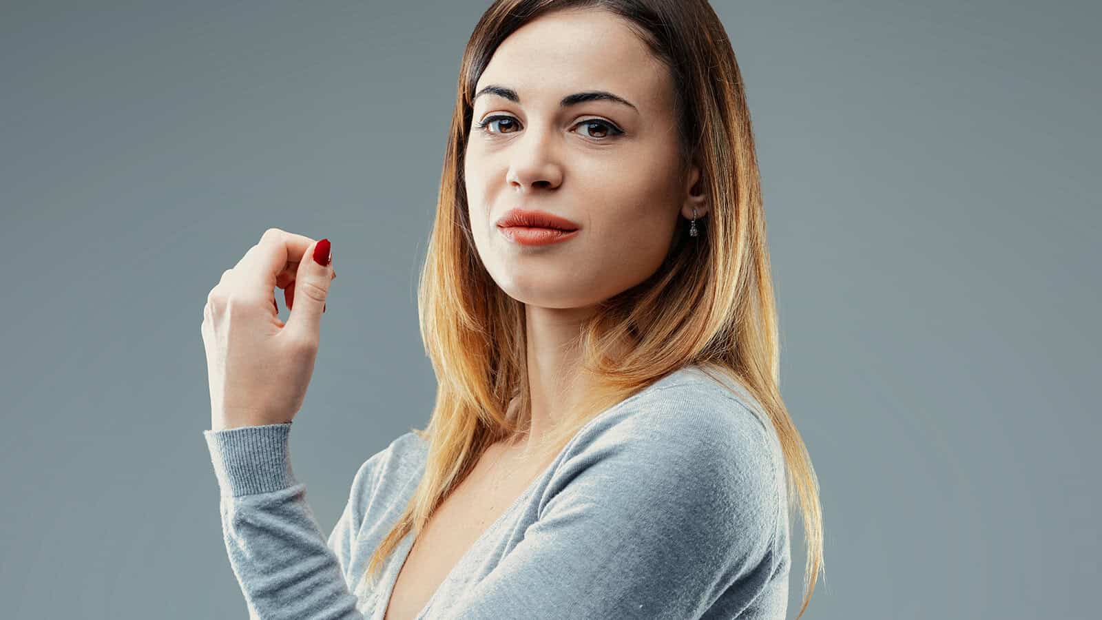 A woman with shoulder-length brown and blonde hair wearing a grey sweater looks toward the camera.
