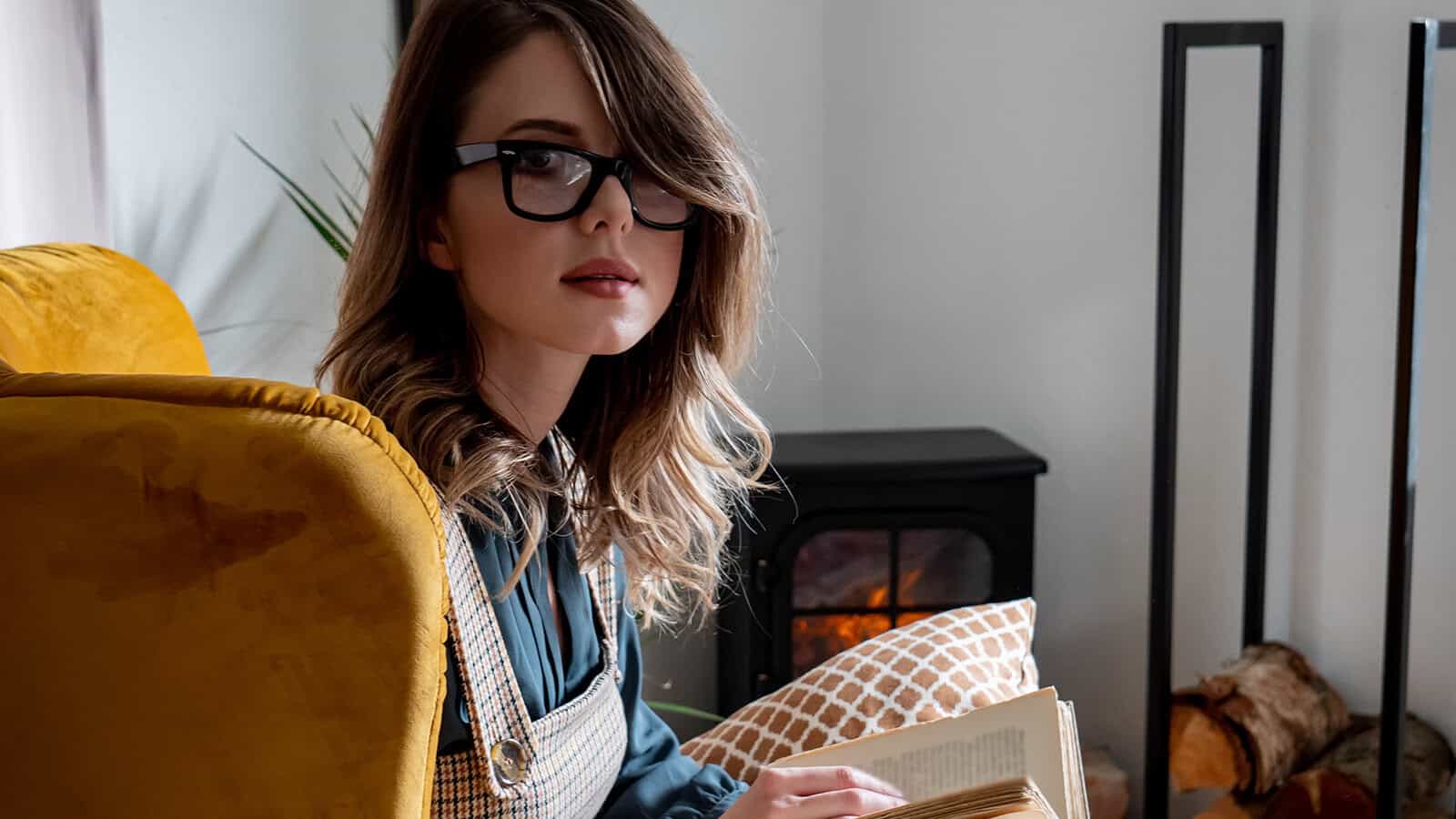 A woman with glasses sitting in a yellow armchair reading a book by a fireplace.