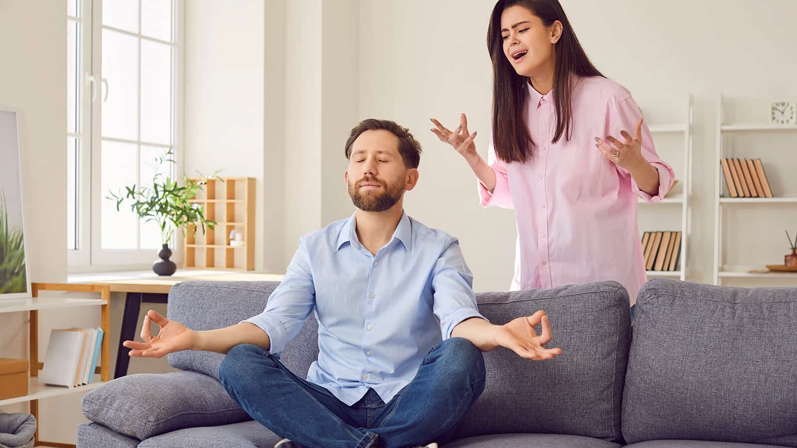 A man sits calmly on a sofa in a meditative pose with his eyes closed while a woman stands behind him, gesturing and looking frustrated.