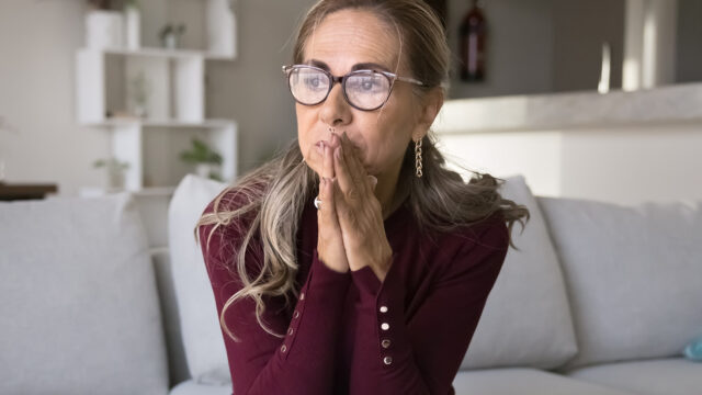 A woman with glasses sitting on a sofa with her hands clasped in front of her face, looking pensive.