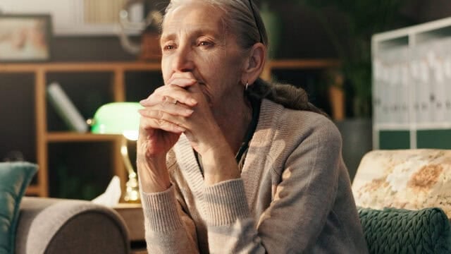 An older woman sits in a living room with her hands clasped, looking away with a thoughtful expression.
