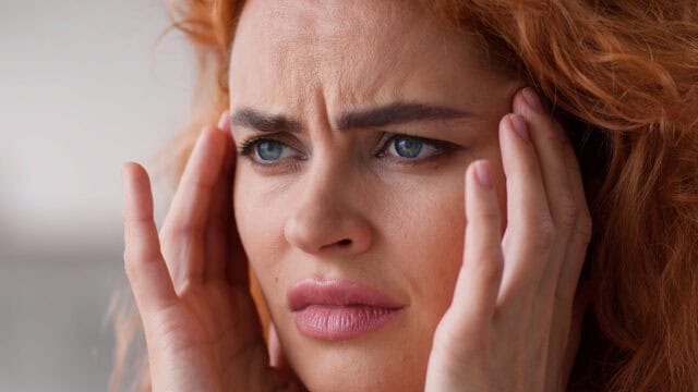 A person with red hair looking distressed while holding their hands to their temples.