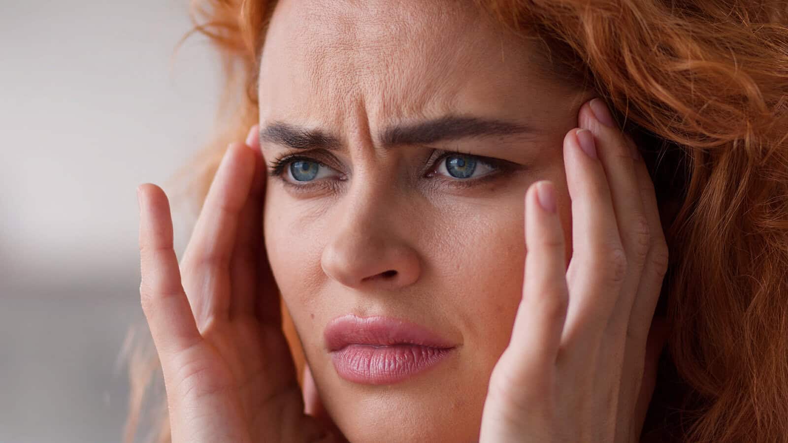 A person with red hair looking distressed while holding their hands to their temples.
