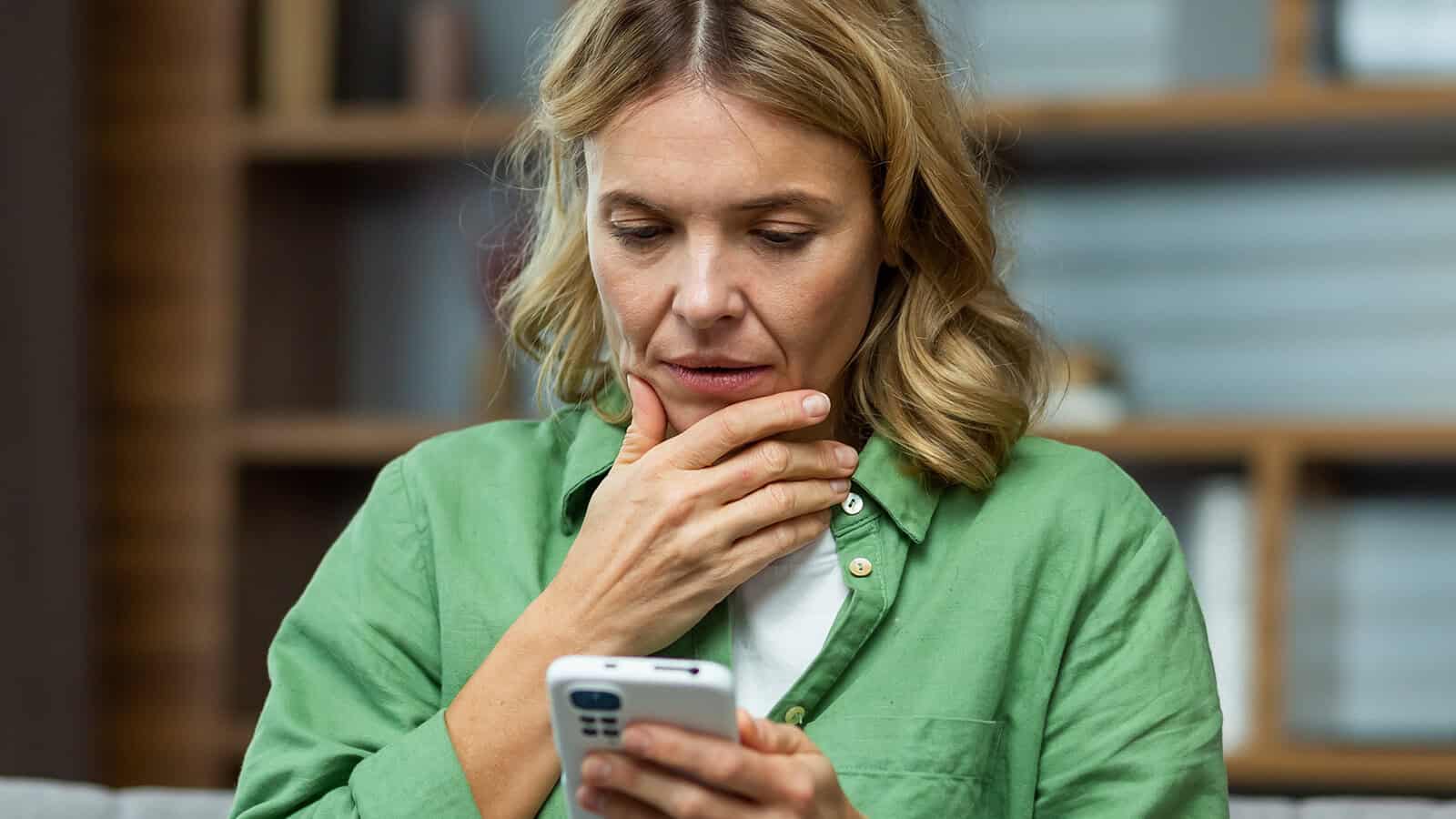 A woman with blonde hair wearing a green shirt looks concerned while holding her phone and touching her chin. She is indoors, with shelves and blurred objects in the background.