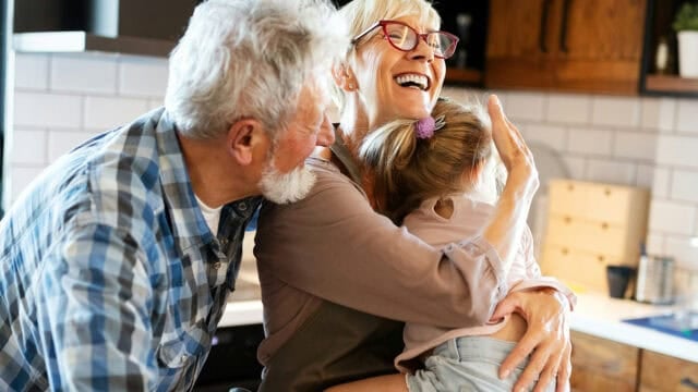 An older couple laughing while hugging a young child in a kitchen.