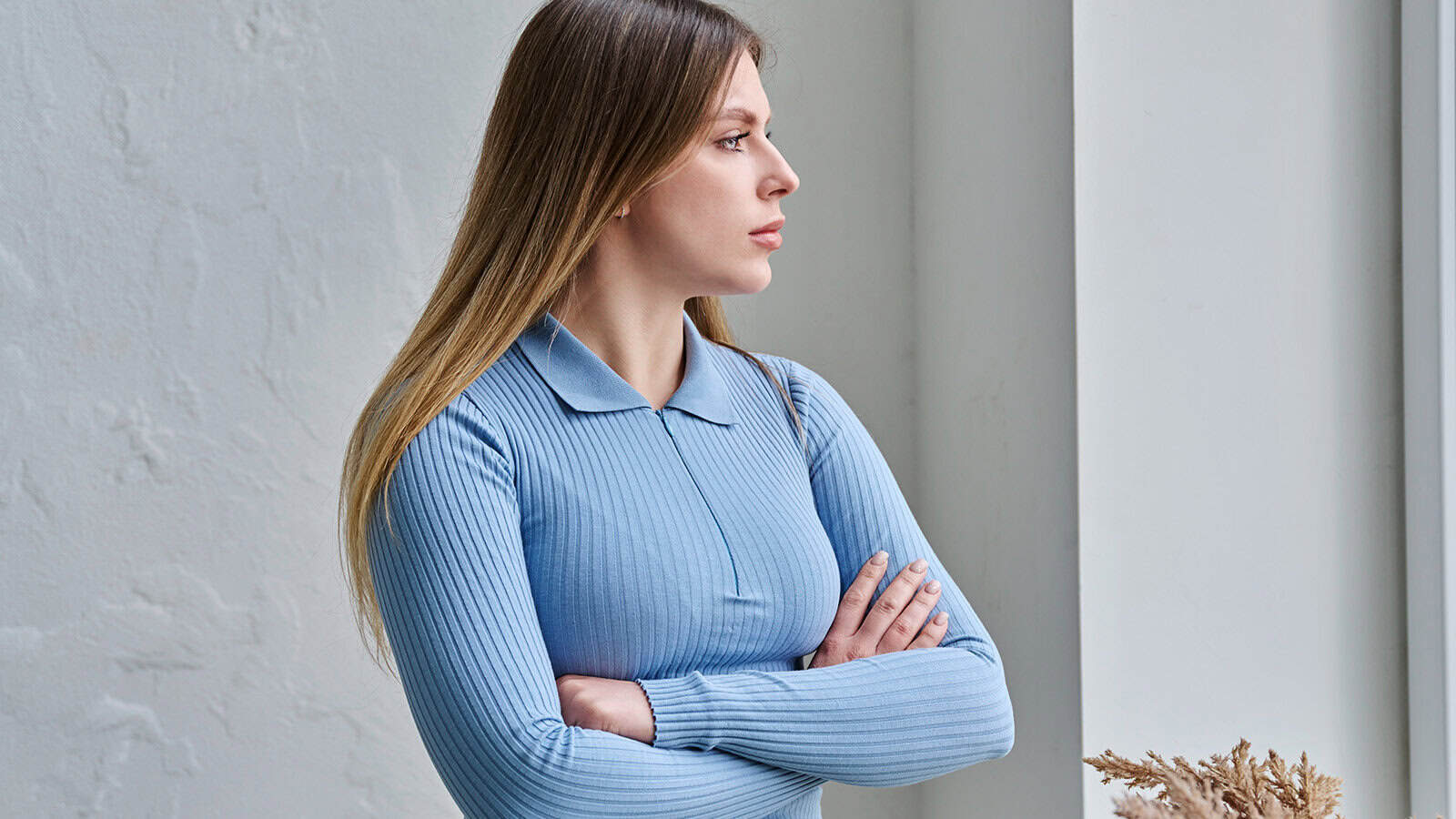 A woman in a blue collared shirt stands with her arms crossed, looking toward a window.