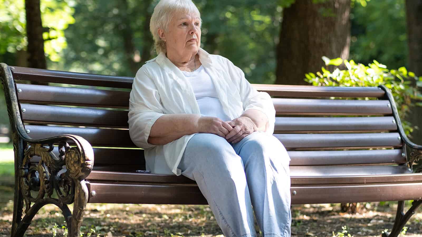 An elderly woman with short gray hair sits alone on a wooden park bench, wearing a white shirt and light blue pants, looking thoughtful. Green trees and sunlight fill the background.