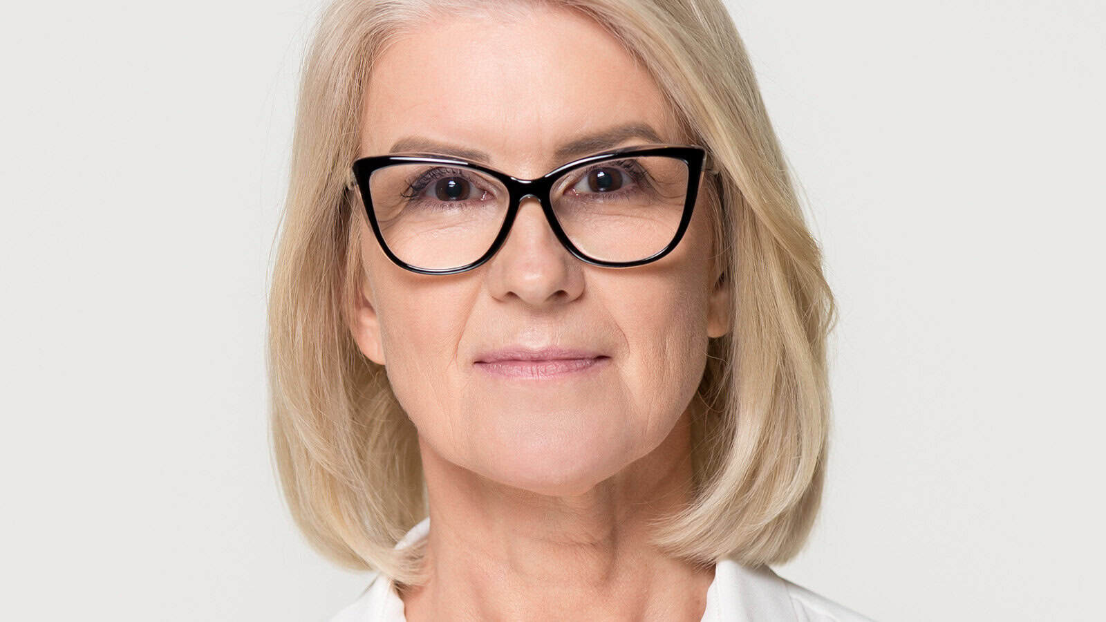 A woman with short blonde hair and black glasses looks forward with a neutral expression, set against a plain light background.