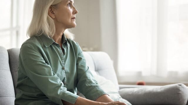 An older woman with grey hair sits on a sofa, looking thoughtfully toward the side.