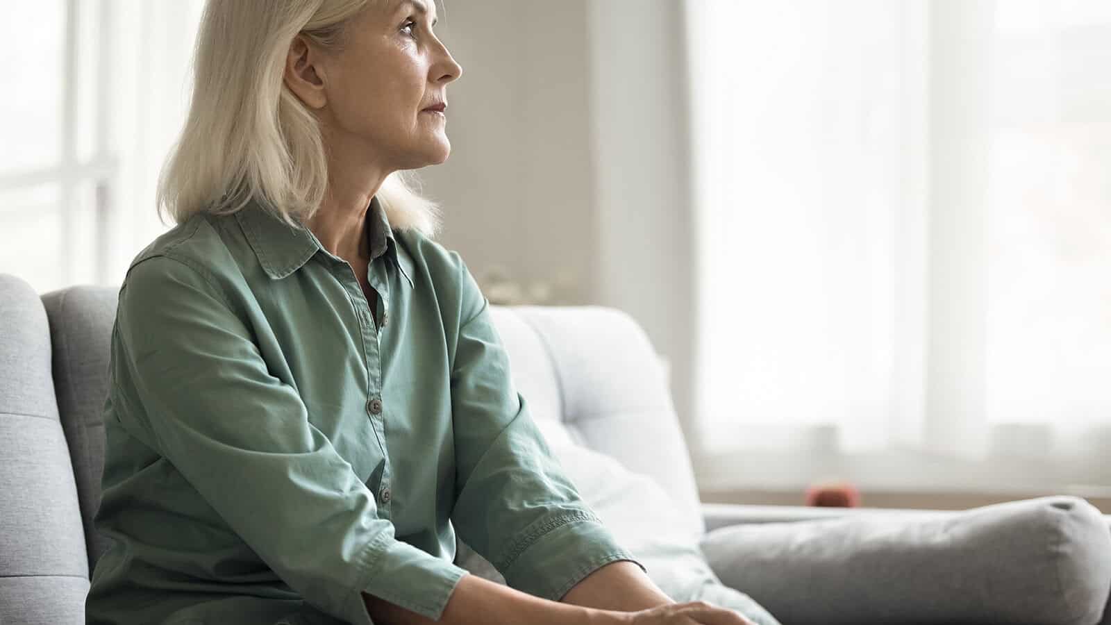 An older woman with grey hair sits on a sofa, looking thoughtfully toward the side.