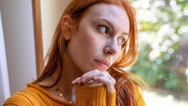 A person with red hair and a yellow sweater rests their chin on their hands while looking away, capturing a moment of quiet loneliness.