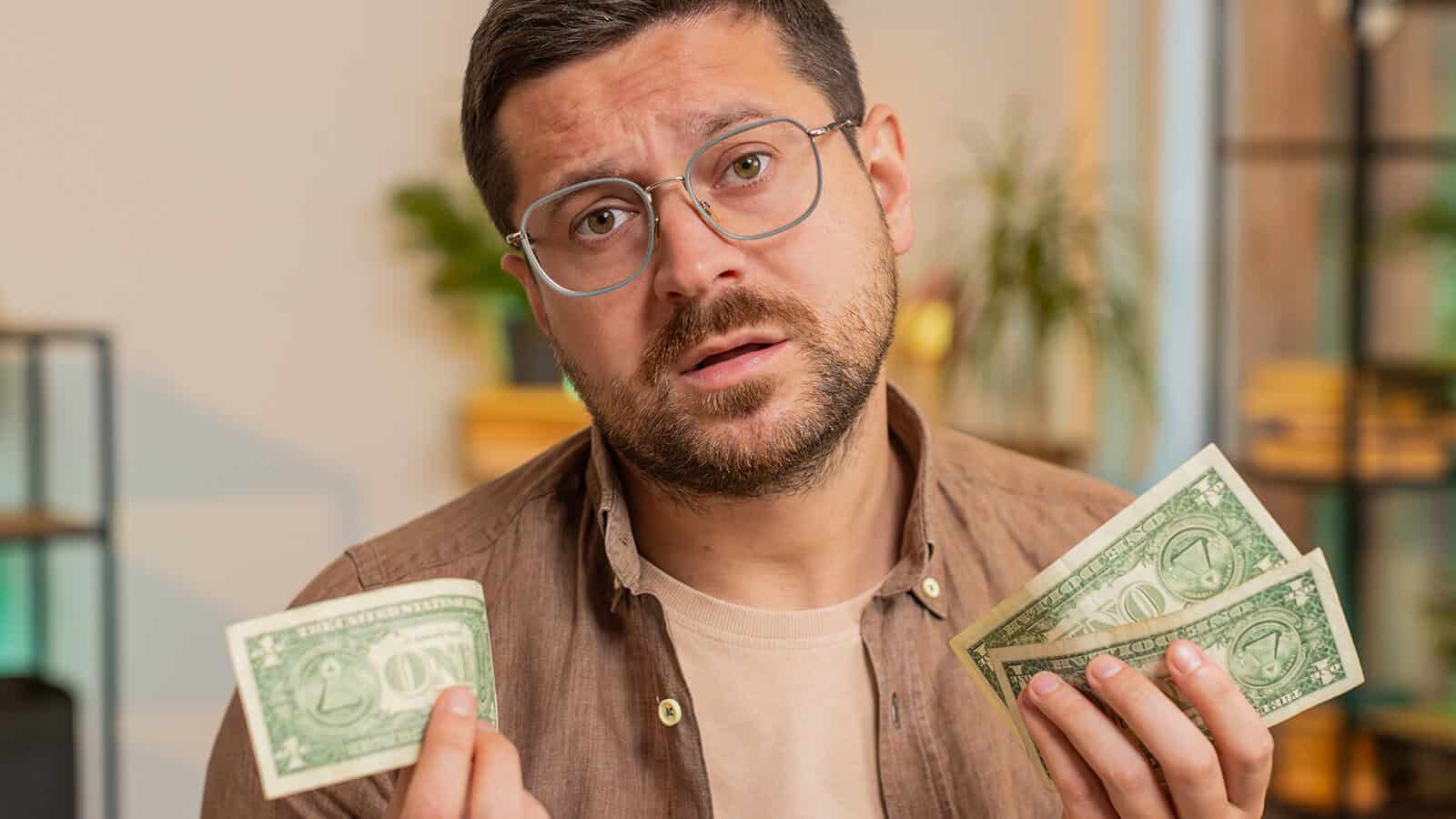 A man with glasses looks skeptically at the camera while holding a few one-dollar bills in his hands.