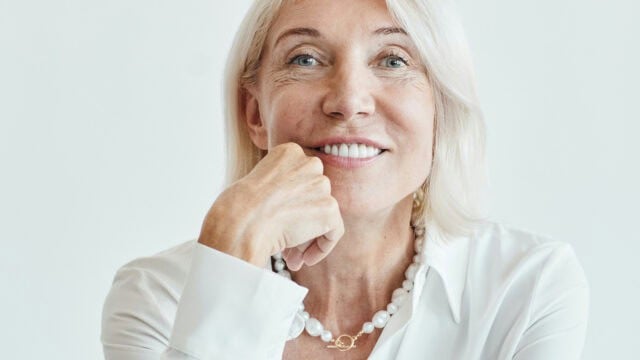 A smiling mature woman with blonde hair wearing a white shirt and a pearl necklace, resting her chin on her hand.