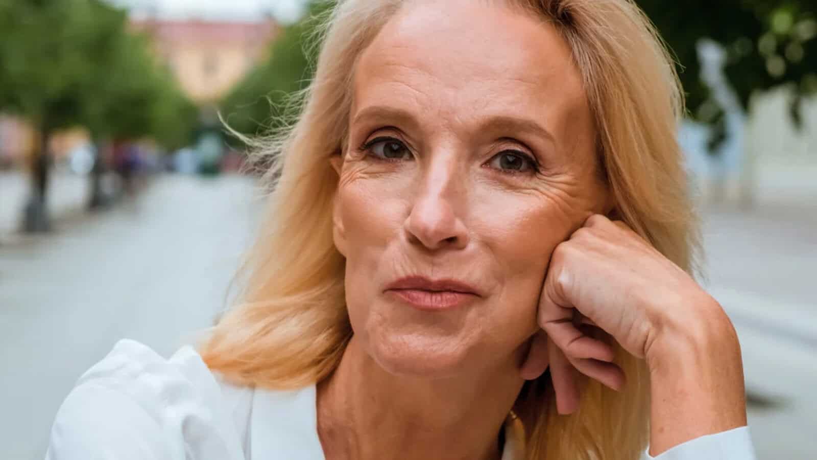 A blonde woman with light skin, wearing a white shirt, rests her head on her hand and looks at the camera with a slight smile while sitting outdoors along a tree-lined street.