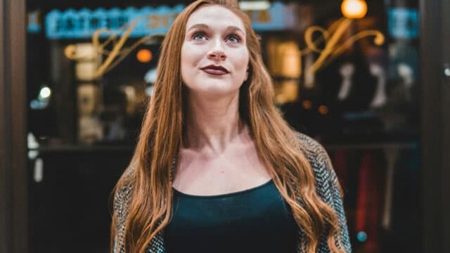 A woman with long red hair and a patterned sweater stands outside, looking upward with a slight smile. Warm lights and window reflections are visible in the blurry background.