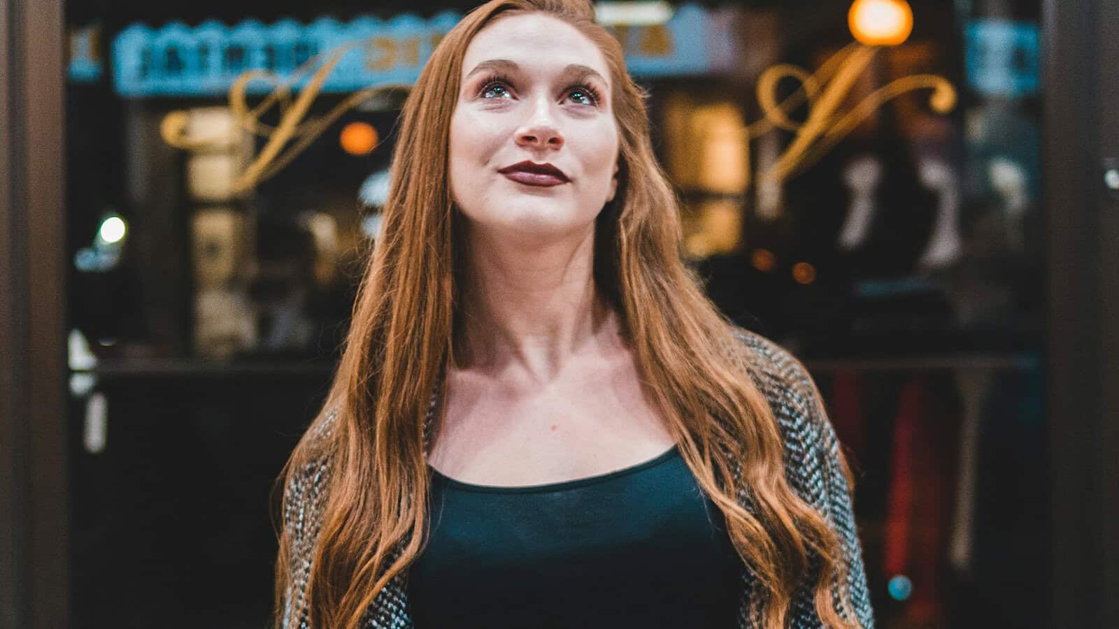 A woman with long red hair and a patterned sweater stands outside, looking upward with a slight smile. Warm lights and window reflections are visible in the blurry background.
