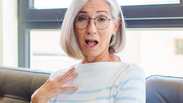 Older woman with gray hair and glasses, sitting on a couch, looking surprised with her mouth open and one hand on her chest.