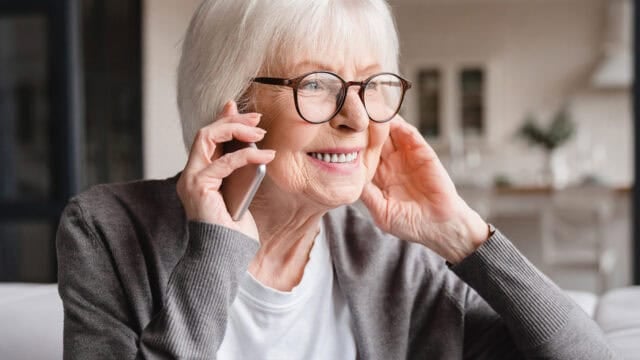 An older woman with white hair and glasses smiles while talking on a smartphone.