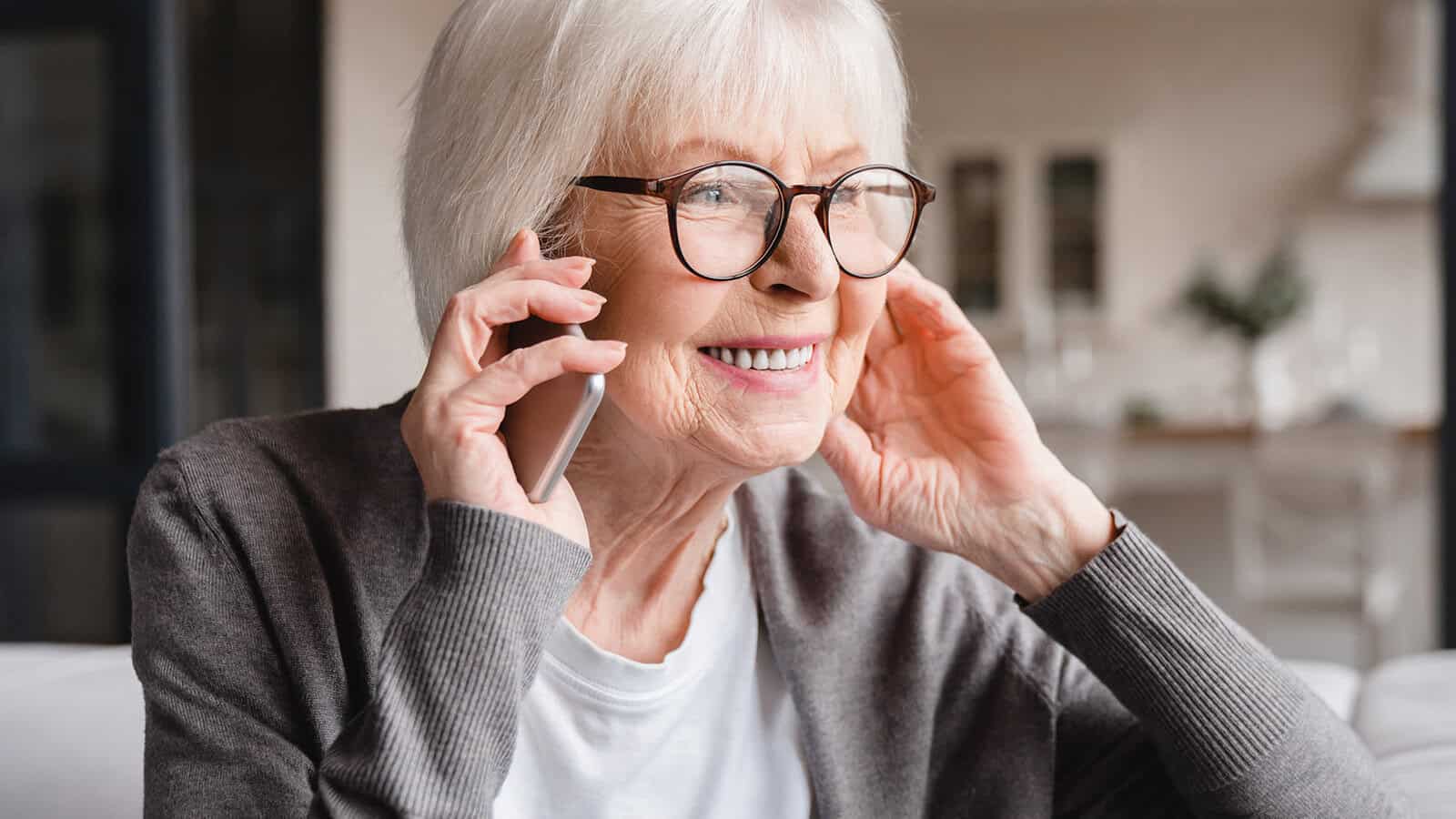 An older woman with white hair and glasses smiles while talking on a smartphone.