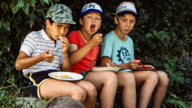 Three children wearing caps sit outdoors eating from plates.