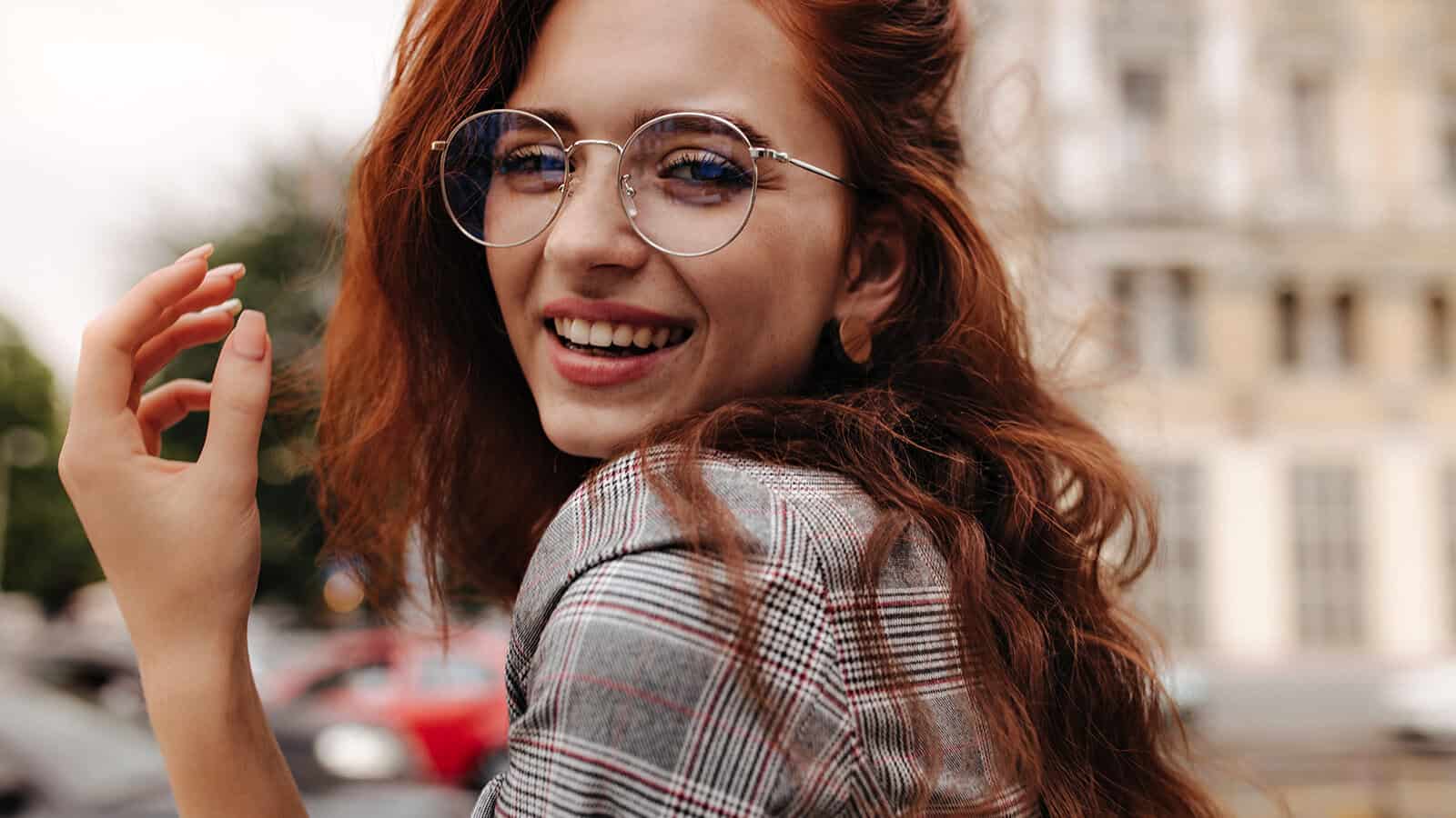 A smiling woman with long red hair wearing round glasses and a plaid blazer looks back over her shoulder.