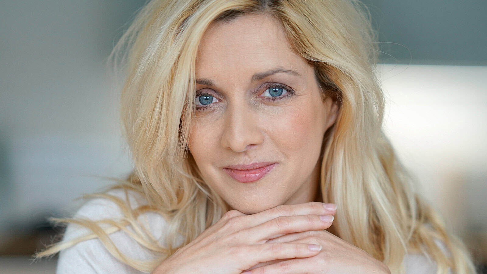 A close-up portrait of a blonde woman resting her chin on her hands and looking at the camera.