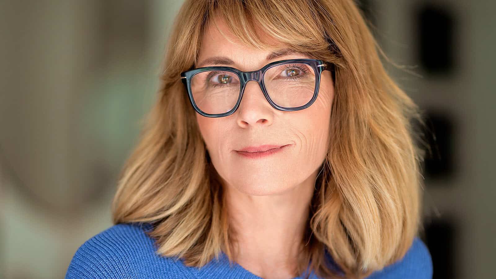 A middle-aged woman wearing black-rimmed glasses and a blue sweater looks directly at the camera.